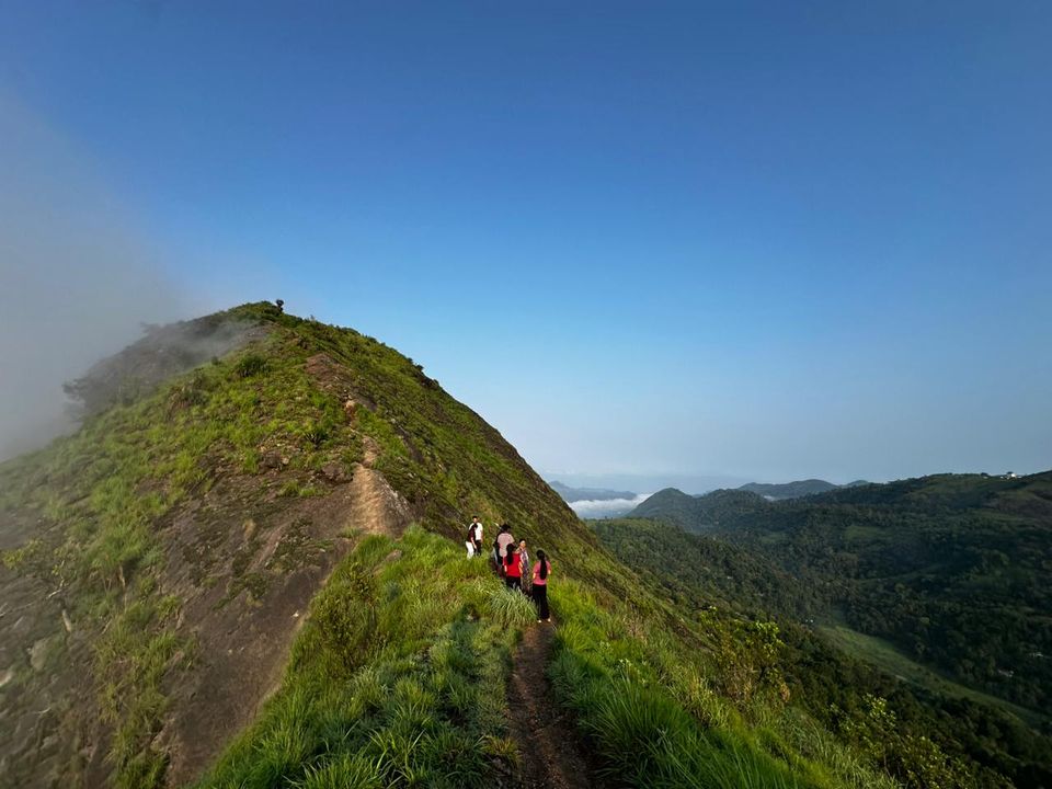 Walkers on a narrow path along a grassy ridge, cloud filling the valley below and mist blowing over the summit